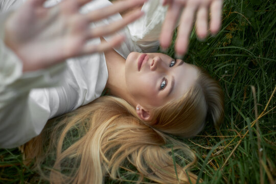 A young woman lies on green grass with her hands raised towards the camera. She has long hair and looks relaxed while enjoying the outdoor scene. Bright light touches her face