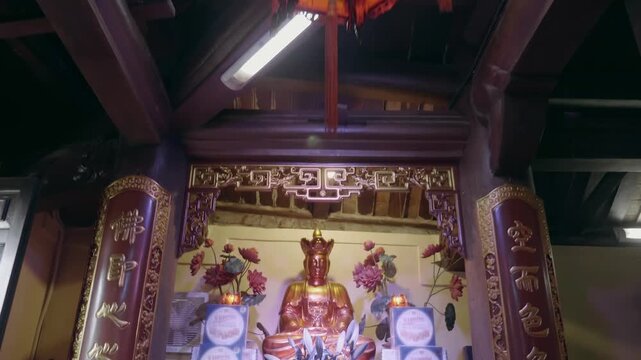 An indoor altar of Temple featuring a central golden seated deity figure (Buddha or a Bodhisattva), adorned with fresh flowers, lilies in a ceramic vase, and stacked apple fruit offerings