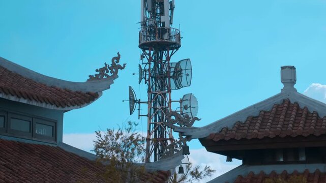 The silhouette of Buddhist Temple with traditional Asian style bended tiled roofs with a large telecommunications and broadcasting lattice tower with multiple antennas that sits on the mountain peak 