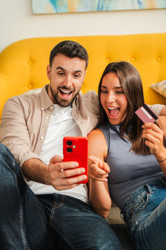 Two smiling partners lounge on a warm sofa, one holds a red smartphone while the other waves a card, creating a lively moment of shared activity and consumer interaction captured candidly.