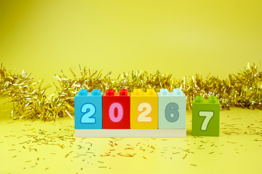 Colorful number blocks showing the transition from 2026 to 2027 with festive gold decoration on a yellow background.