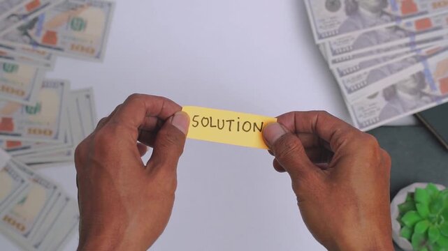 Person holding a yellow sticky note with the word SOLUTION written on it surrounded by stacks of hundred-dollar bills.