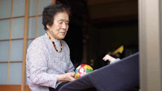 Older woman sits near a baby bouncer and holds a colorful patchwork ball in both hands.