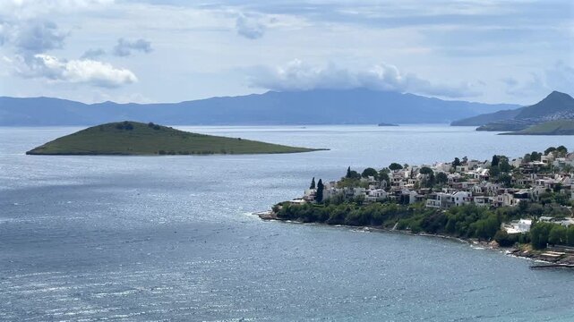 A high-angle view of white houses on a green peninsula jutting into the blue sea, with a small island and cloud-capped mountains in Bitez, Turkey.
