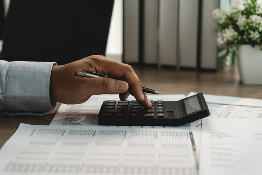 Businessman using calculator on financial documents while holding pen, showing financial analysis, accounting review, budget calculation and business reporting workflow.