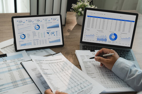 Financial data review on laptop spreadsheet and tablet dashboard with printed reports and pen, showing accounting analysis and business finance workflow.