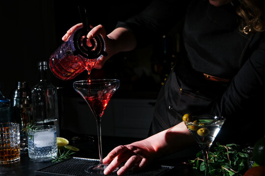 Bartender pouring red cosmopolitan cocktail drink into martini glass at bar counter. Modern mixology craft and hospitality business. Professional preparing cold aperitif beverage for evening party.