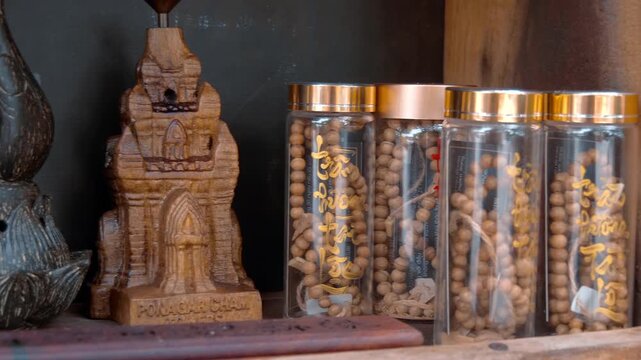 A carved wooden miniature model of the historic Po Nagar Cham Temple Tower next to clear glass jars with golden lids and Vietnamese characters filled with wooden prayer beads. Shot in motion