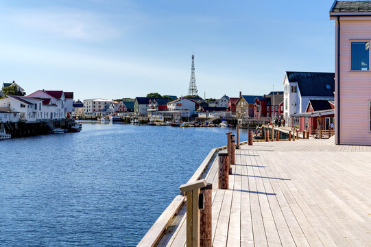 Sunny harbor view of Henningsv&aelig;r on the Lofoten Islands with traditional wooden houses, boats, calm water and a wide waterfront promenade under blue sky on a clear autumn day.