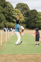 Obraz premium Adult man playing cricket, releasing the ball with a classic bowling action on a grassy pitch while a young child watches and other people gather in the background