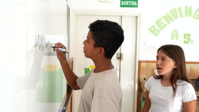 Focused preteen boy writing on a whiteboard with a marker during a lesson, with his curious female classmate watching and interacting next to him in a bright elementary school classroom