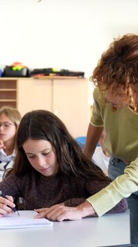 Middle aged female teacher with curly hair helping a young girl with her schoolwork, guiding her writing in a notebook while she sits at her desk in a bright elementary school classroom