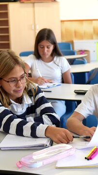 Young students sitting at their desks during a class lecture, secretly passing a folded note to each other while trying not to get caught, representing a classic school life moment