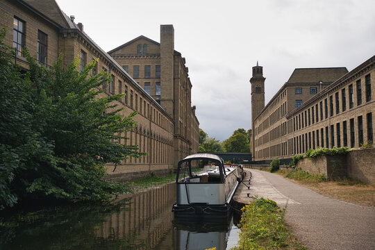 Long canal boat navigating calm water, reflecting old industrial architecture of a traditional brick mill building with its prominent tower under a cloudy sky, showcasing england's heritage