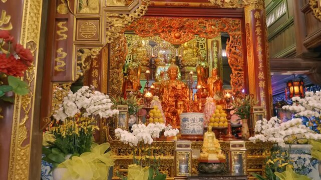 Traditional Buddhist altar adorned with golden Buddha statues, glowing lamps, offerings of fruit, and white orchids. Golden Buddha figure sits at the center, surrounded by smaller attendant figures 
