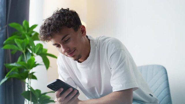 Young man with curly hair texting and smiling at home