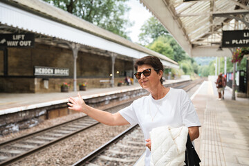 Mature woman in sunglasses and white t shirt stands on a historic train platform in hebden bridge, smiling and extending her arm while waiting for a train, summer daylight ambiance © Fotografia Juan Reig