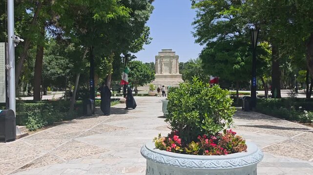 Women in black chador pass Ferdowsi Tomb beneath Iranian flags in a public heritage site.