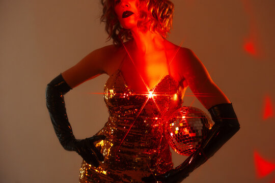 Close-up of gold sequin dress and mirror ball in red light, woman with dark lips, cropped face