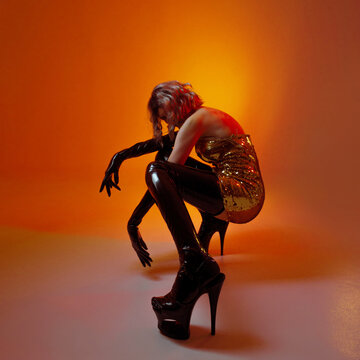 Faceless fashion figure in gold sequins and black vinyl crouching, head bowed, orange glow