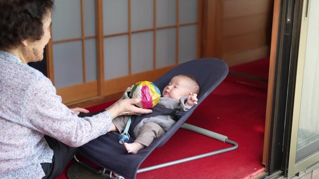 Older woman reaches a colorful patchwork ball toward a baby reclining in a blue bouncer.