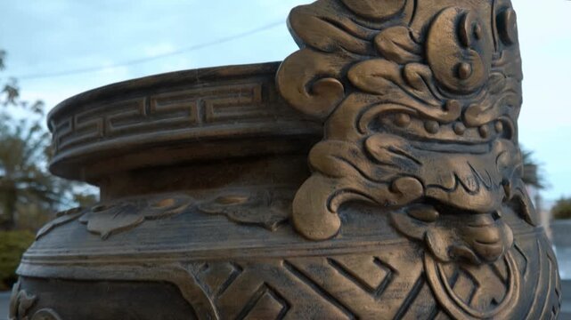 A close-up of a massive bronze vase or urn for incense offering carved with dragon and floral patterns placed near the feet of the giant white stone Buddha statue on the hill in Long Son Pagoda 