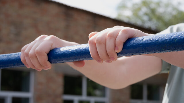 Close-up of a child's hands gripping a blue metal horizontal pull-up bar outdoors. Active kid practicing pull-ups on sports playground with blurred background and sunny daytime lighting.