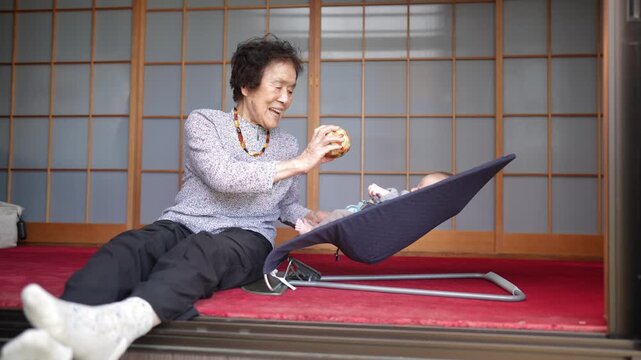 Older woman smiles while holding a colorful patchwork ball beside a baby reclining in a bouncer.