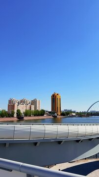Pedestrian bridge over Ishim River in Astana, Kazakhstan, with city architecture, blue water and open summer sky for travel stock footage, Kazakhstan, shot handheld, vertical video, 4k 