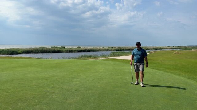 Male golfer walks across coastal putting green carrying a golf club beside water, sand bunker, marsh grass, and ocean under cloudy skies in scenic outdoor course