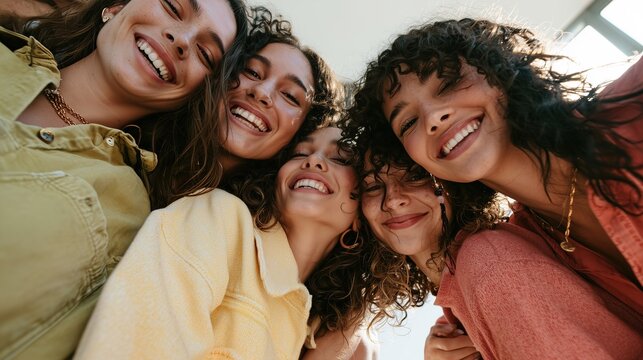 Low angle view of six diverse young women smiling broadly and looking at camera together, huddling closely. Their faces are joyful and filled with genuine laughter, while their varied textured hair fr