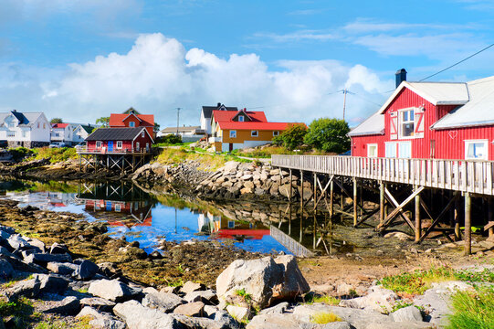 Traditional red and white wooden houses stand beside a small rocky bay in Henningsv&aelig;r on the Lofoten Islands, while buildings reflect in calm water on a sunny autumn day.