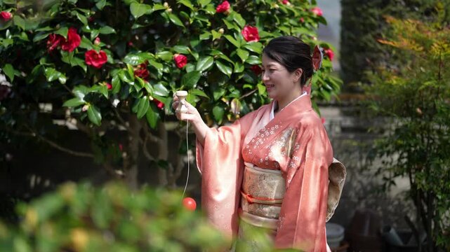 Adult woman in an orange kimono raises a red ball attached to a string outdoors beside a flowering shrub.