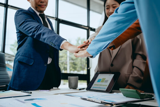 Diverse business team stacking hands together over a desk with charts and tablet in a bright modern office, showing teamwork and partnership