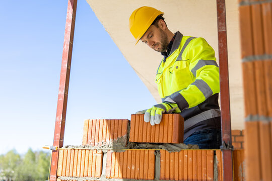 Skilled builder wearing safety helmet positioning red brick block on mortar