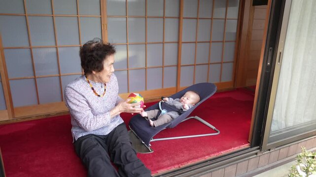 Older woman turns toward a baby in a bouncer while holding a colorful patchwork ball in both hands.