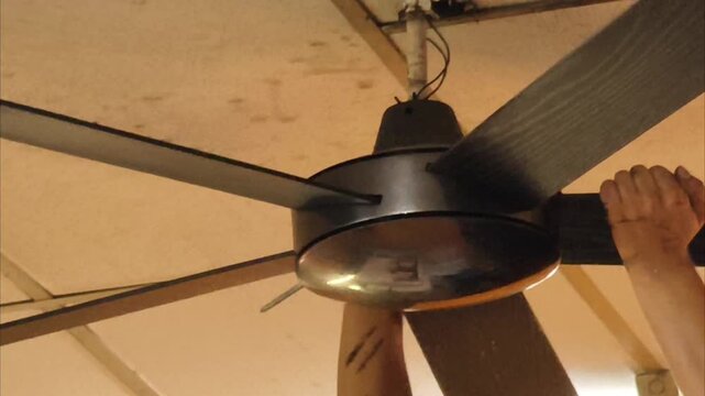 ​A technician is carefully installing and adjusting a dark metal ceiling fan with five blades on an indoor ceiling using a screwdriver.
