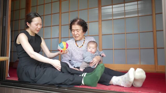 Two adult women sit together at an open doorway with a baby and a colorful patchwork ball.