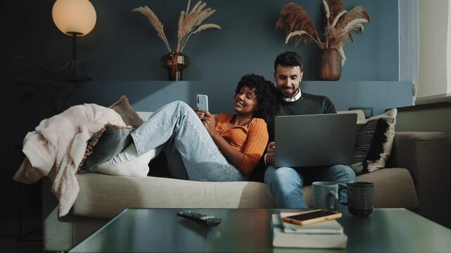 Couple on sofa enjoying, social media and streaming subscription, using laptop and mobile phone. African American woman and caucasian man