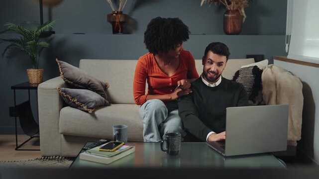 Happy multiracial couple sitting on the sofa and making an online payment using credit card and laptop