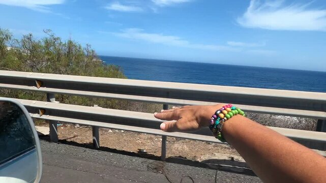An adventurous journey unfolds as a hand, adorned with colorful wooden bracelets, reaches out from a moving car, playfully catching the invigorating ocean breeze along a scenic coastal highway.