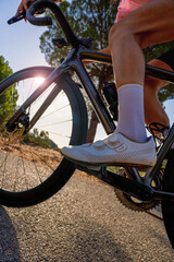 A low-angle macro close-up of a female cyclist's shoe and pedal in motion as she climbs a mountain pass on the Costa Brava on her road bike, with sunbeams filtering through the wheel spokes.
