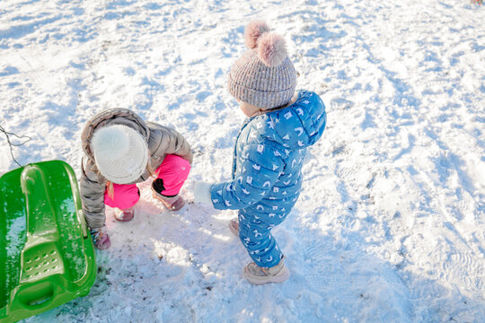Two little girls playing together in deep snow with green plastic sled during sunny winter outdoors activity, children enjoying snow games in countryside meadow, active kids friendship and winter vaca
