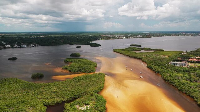 Rust-colored tidal channel winds between mangrove islands as recreational boats cruise toward deeper dark water on a humid day.
