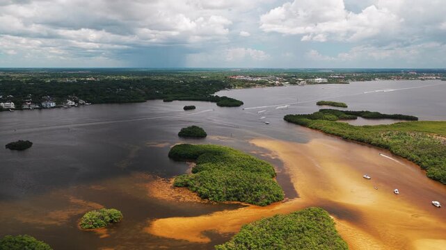 Motorboats weave through a brown estuary past bright sandbars and mangrove patches as a rainstorm builds on the distant horizon.