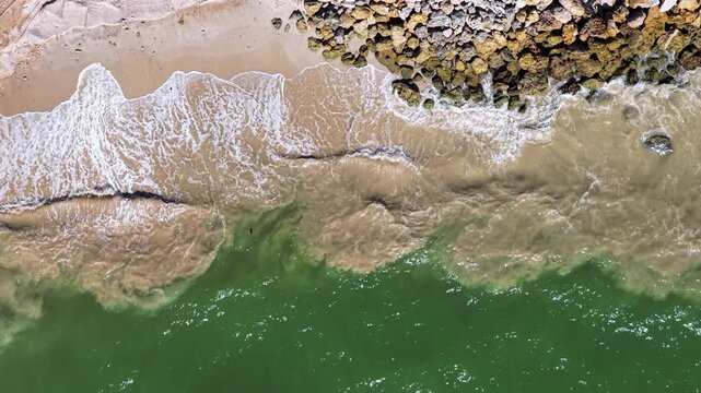 Foamy waves roll over brown sand and crash along a rocky breakwater as the drone hovers above the shoreline.