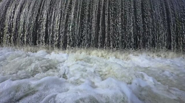 Close up of rushing water curtain overflowing from river weir dam into foamy riverbed