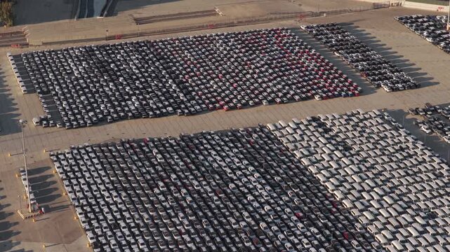 Aerial perspective showing thousands of new cars neatly parked in a vast lot at a port terminal, waiting for distribution and shipment for the automotive industry during sunset