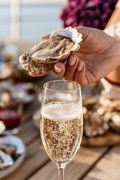 A person's hand holding an open oyster in its shell over a wooden table. In the foreground, a glass of sparkling white wine or champagne.