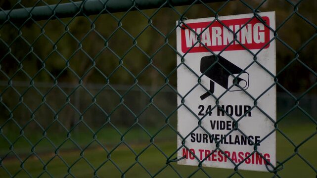 Slow motion close-up of a metal warning 24 hour video surveillance sign with no trespassing security lettering and a camera icon attached securely to a chain-link fence gate for protection Australia